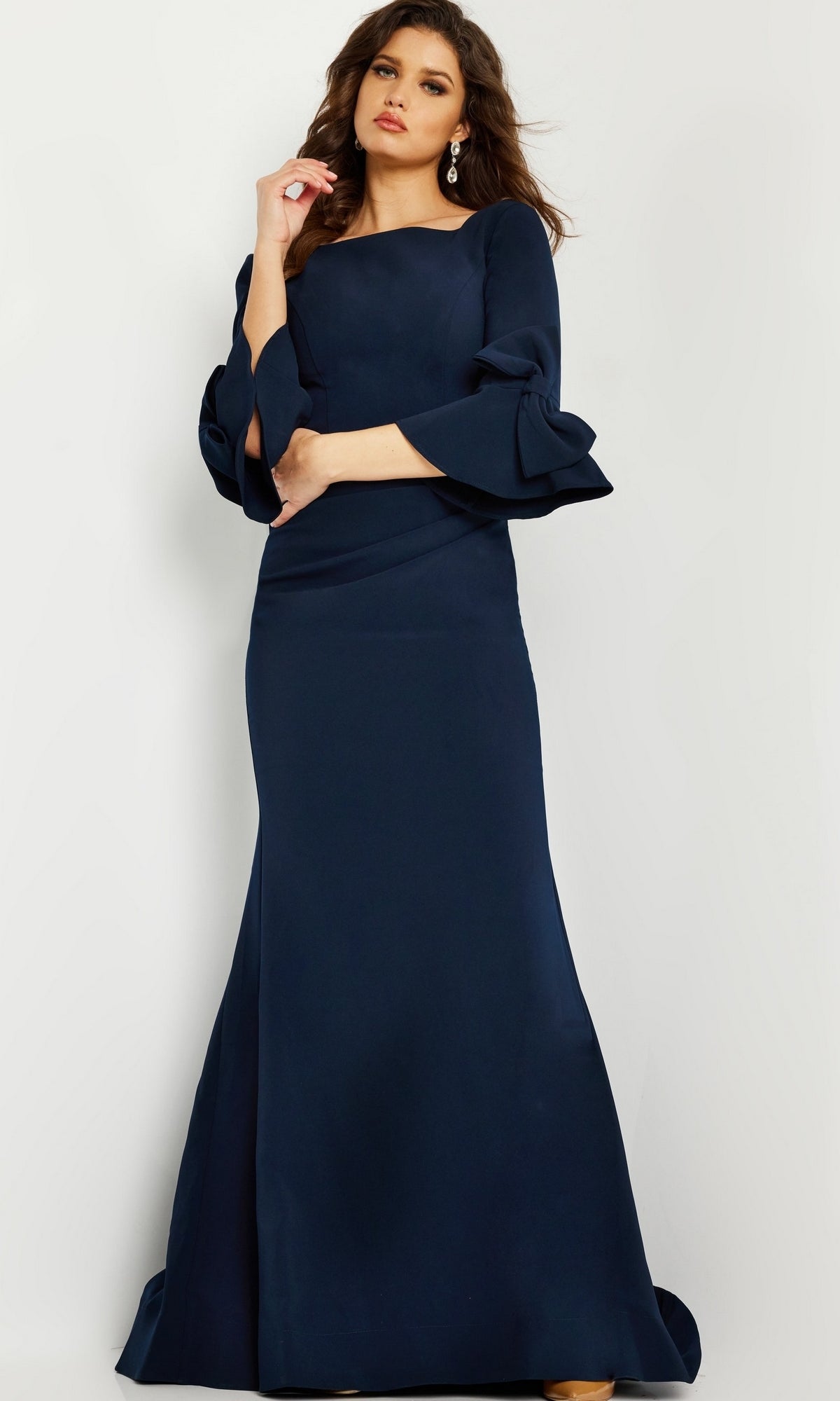 A woman stands against a light background wearing the Long Formal Jovani Dress 09776, a navy blue evening gown with three-quarter bell sleeves.